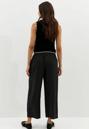 Kobieta z długimi brązowymi włosami stoi tyłem, ubrana w czarny bezrękawnik z białą lamówką, luźne czarne spodnie i czarne buty wsuwane.