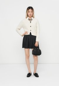 Cárdigan de punto trenzado color crema con botones negros, combinado con una falda negra con pliegues y un bolso negro con un diseño texturizado. Unos mocasines negros complementan el conjunto.