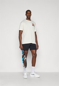 Koszulka z krótkim rękawem w kolorze kremowym z logo, czarne szorty, białe trampki, trzymająca rakietę tenisową, stojąca na jednolitym tle.