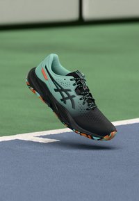 Fekete és türkiz sportcipő, légáteresztő hálós felsőrésszel, textúrázott részletekkel és sokszínű mintázatú talppal, pályasportokhoz tervezve.