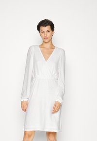 Vestido branco com decote em V, mangas bufantes longas e tecido texturizado. Apresenta uma cintura elástica e punhos franzidos para um ajuste confortável.