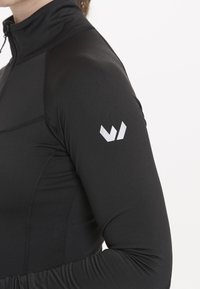 Sort langærmet atletisk top med halv-lynlås design. Har en glat tekstur og et reflekterende hvidt logo på højre skulder.