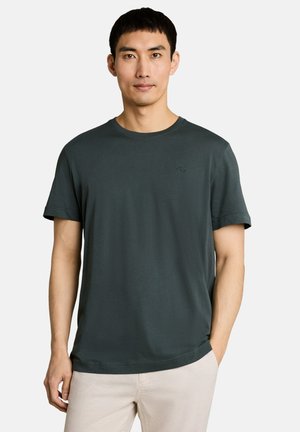 Jonge man met kort zwart haar, die een donkergroen T-shirt met korte mouwen en lichtbeige broek draagt, staand tegen een effen achtergrond.