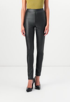Donna che indossa pantaloni in pelle neri a vita alta e tacchi alti neri, in piedi davanti a uno sfondo bianco semplice.