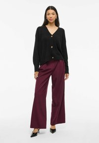 Černý žebrovaný cardigan s výstřihem do V a zlatými knoflíky, v kombinaci s širokými burgundskými kalhotami a černými špičatými podpatky. Modelka na neutrálním pozadí.
