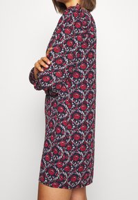 Donna in un vestito blu navy a maniche lunghe con motivo floreale rosso, in piedi di lato con le braccia incrociate su uno sfondo bianco uniforme.