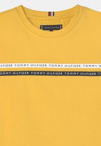 Żółta bawełniana koszulka z okrągłym dekoltem. Posiada poziomy pasek z napisem "TOMMY HILFIGER" w czerni i bieli. Gładka faktura.