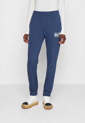 Persoon draagt marineblauwe joggers met elastische enkels en een wit logo op de dij, gecombineerd met witte en zwarte sneakers met een captoe.