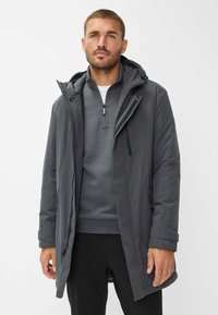 Uomo che indossa un giaccone invernale grigio con cappuccio sopra un maglione grigio con zip e pantaloni neri, in piedi di fronte a uno sfondo bianco semplice.