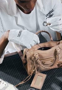 Personne portant des gants blancs inspecte un sac à main en cuir beige à l’aide d’une loupe et d’un outil métallique.