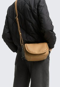 Braune Leder-Crossbody-Tasche mit einer strukturierten Oberfläche und einem gebogenen Deckel, ausgestattet mit einem langen Riemen, der mit einem schwarz-weißen Quastenakzent verziert ist.