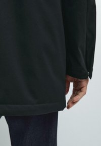 Schwarze Softshelljacke mit glatter Textur, gerader Schnitt und Knopfdetails an den Manschetten, kombiniert mit dunklen Jeans.