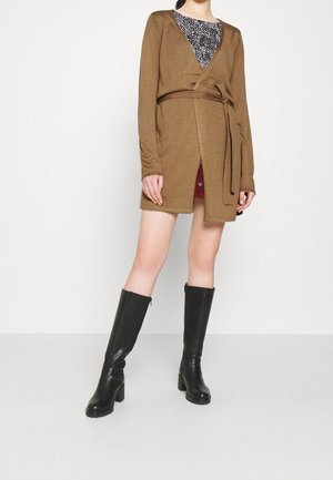 Femme portant un cardigan marron ceinturé par-dessus un haut à motifs noir et blanc, associé à des bottes noires montantes jusqu'aux genoux.
