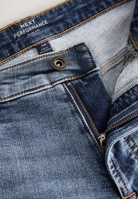 Niebieskie dżinsy o dopasowanym kroju z częściowo rozpiętym zamkiem błyskawicznym, metalowym guzikiem oraz widoczną wewnętrzną metką z rozmiarem 34R, wyprodukowane w Bangladeszu.