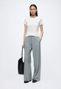Mujer está de pie con una camisa blanca de manga corta y pantalones anchos gris, sosteniendo una bolsa negra con una mano en el bolsillo.