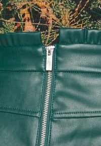 Grön läderplagg med dragkedjestängning, med textilerad kamouflage överdel i olika jordnyanser som framhäver en blandad materialdesign.