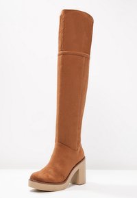 Botte cuissarde en daim marron avec une semelle plateforme épaisse. Présente une texture lisse et un talon large et structuré.