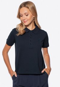 Námořnická modrá polo trika vyrobená z ribbené tkaniny, má límec, čtyřknoflíkovou légu a vyšitý logo na hrudi. Krátké rukávy.