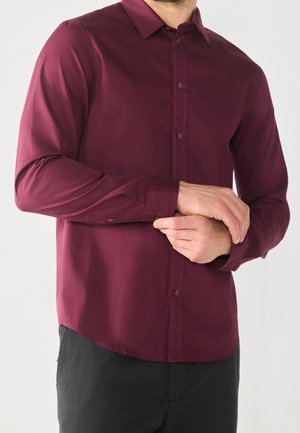 Man die een bordeauxrode, lange mouwen overhemd met knopen draagt en de manchet aan zijn linker pols afstelt, gekleed in een zwarte broek.