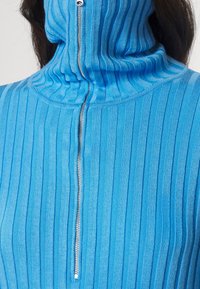 Niebieski sweter z prążkowanej dzianiny z wysokim kołnierzem i przednim zamkiem błyskawicznym. Faktura jest dziana, z pionowymi paskami dla dodatkowego efektu.