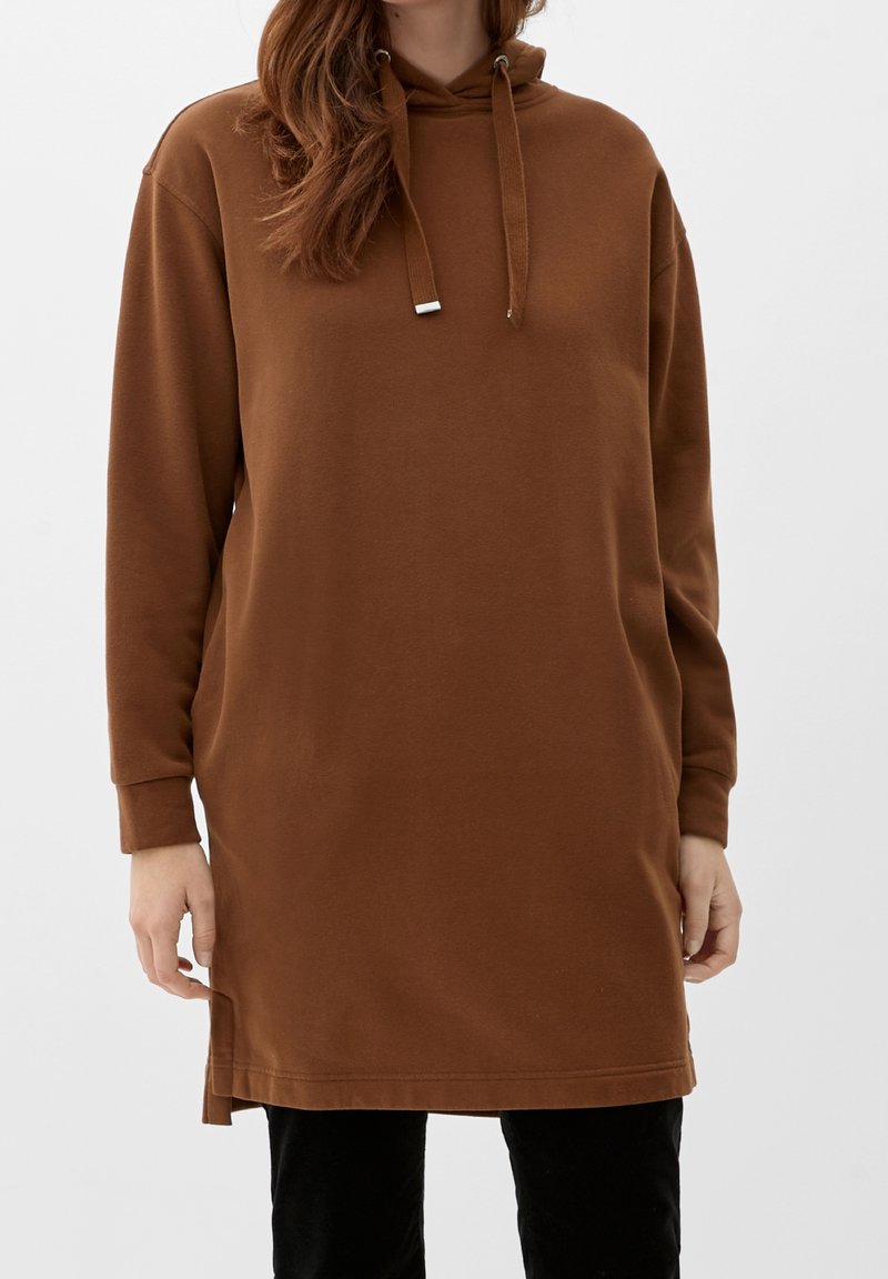 Donna che indossa una felpa con cappuccio lunga marrone con coulisse, abbinata a pantaloni neri, in piedi contro uno sfondo semplice.