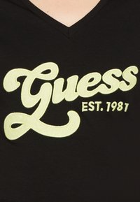 Sort V-hals t-shirt med oversized, broderet gul skrift, der står "Guess EST. 1987." Blødt stof med en glat tekstur.