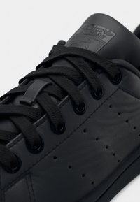 Černá kožená teniska s černými tkaničkami, kulatými průchodkami a perforovaným bočním panelem, s viditelnou značkovou štítkem na jazyku.