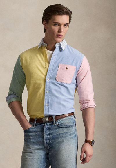 Junger Mann trägt ein mehrfarbiges Hemd mit gelben, blauen, pinken und grünen Einsätzen, in blaue Jeans mit braunem Gürtel gesteckt.