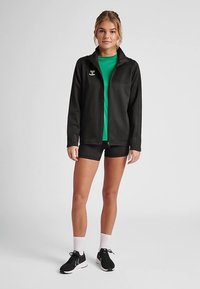 Schwarze Reißverschlussjacke mit hohem Kragen, kombiniert mit einem grünen T-Shirt und schwarzen Shorts. Dazu schwarze Sneakers und weiße Sportsocken.