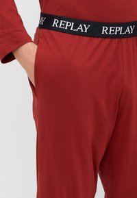 Sarkanas kokvilnas bikses ar melnu elastīgu jostu, kurā redzams "REPLAY" logo. Redzama sānu kabata, gluda tekstūra un brīvs griezums.