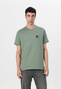 Tricou din bumbac verde cu decolteu rotund și mâneci scurte, prezentând o mică etichetă cu logo negru pe pieptul stâng. Textură simplă, netedă.