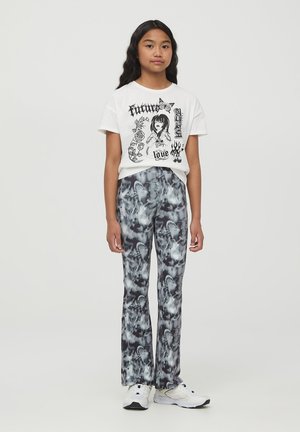 Maglietta grafica bianca con stampa nera che raffigura una figura e del testo. Pantaloni a zampa di elefante in tie-dye bianco e nero con una texture liscia. Sneakers bianche.