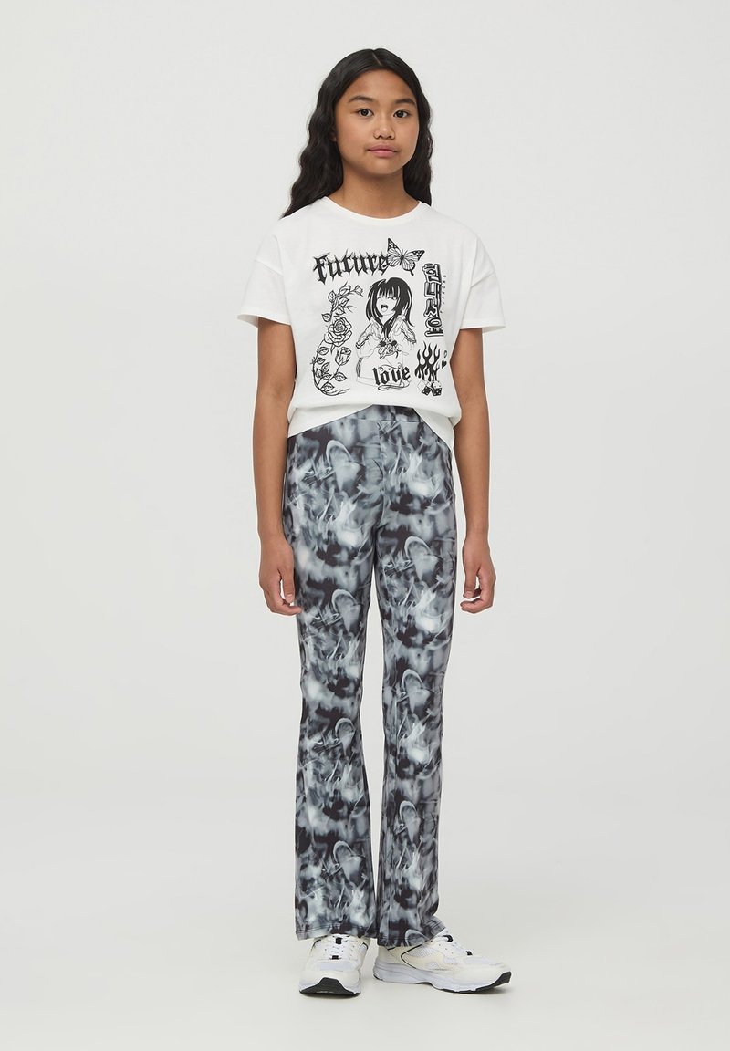 Maglietta grafica bianca con stampa nera che raffigura una figura e del testo. Pantaloni a zampa di elefante in tie-dye bianco e nero con una texture liscia. Sneakers bianche.