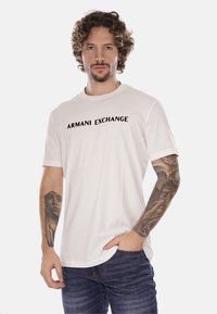T-shirt bianca in cotone con testo nero "ARMANI EXCHANGE" sulla parte anteriore, maniche corte, vestibilità comoda, abbinata a jeans blu scuro.