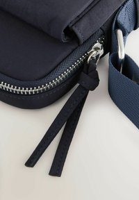 Borsa a tracolla blu navy con un esterno liscio, tracolla testurizzata, zip in metallo argentato e etichette in tessuto sul cursore della zip.
