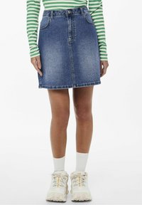 Nainen, jolla on päällään sininen farkkuhame, vihreä-valkoraidallinen pitkähihaisen paita, valkoiset sukat ja valkoiset kömpelöt lenkkarit, seisoo valkoista taustaa vasten.