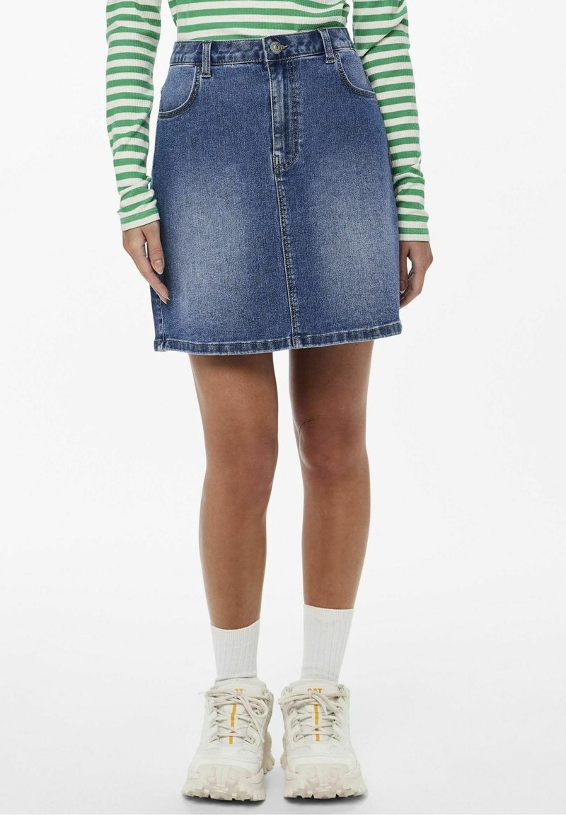 Nainen, jolla on päällään sininen farkkuhame, vihreä-valkoraidallinen pitkähihaisen paita, valkoiset sukat ja valkoiset kömpelöt lenkkarit, seisoo valkoista taustaa vasten.