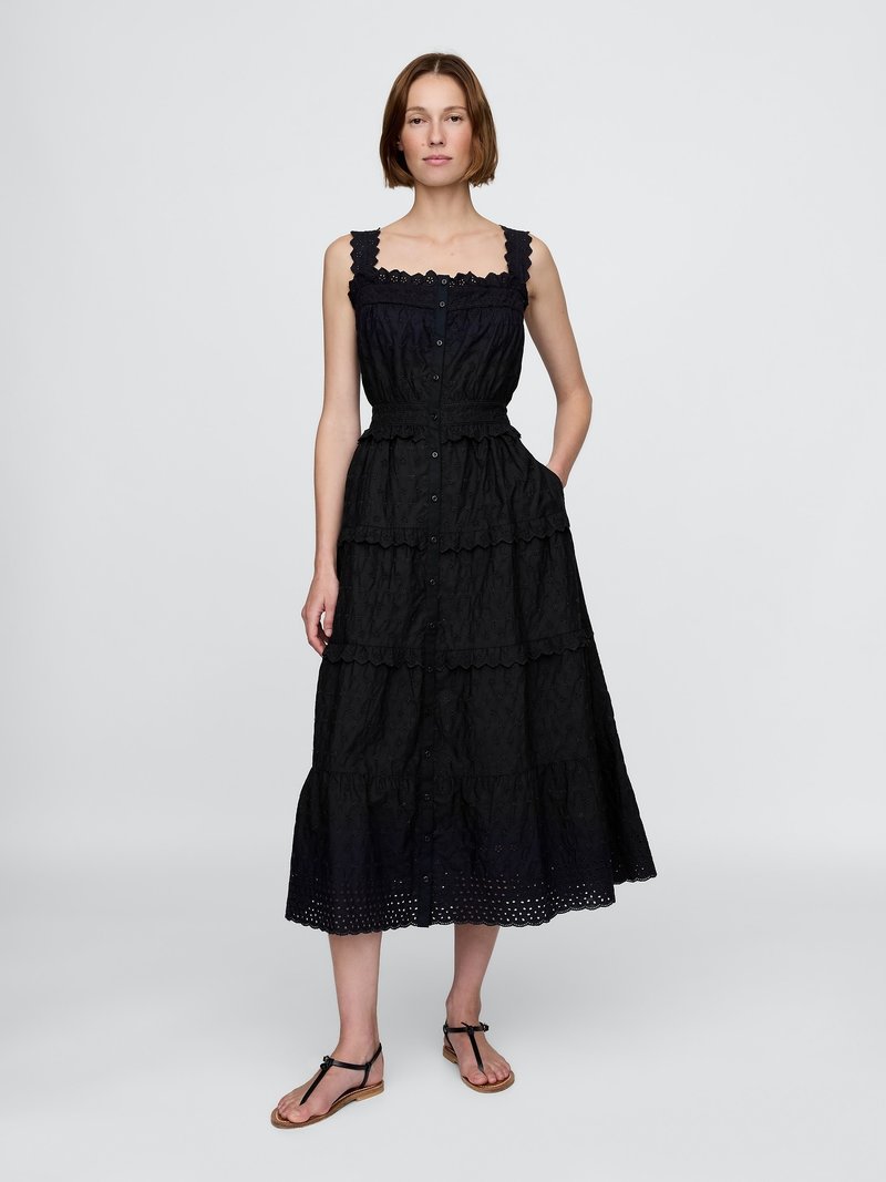 Vestido midi negro con volantes y tirantes de encaje, con botones en la parte delantera y una tela texturizada que presenta patrones florales. Llevado con sandalias planas.