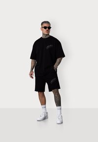 Ensemble comprenant un t-shirt surdimensionné noir et un short en coton, avec des accents brodés. Des baskets blanches et des lunettes de soleil complètent le look.