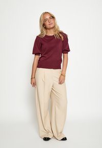 Bordeaux kortemouwtop met een gestructureerde afwerking, gecombineerd met een crèmekleurige broek met wijdere pijpen. Model staat zelfverzekerd tegen een effen achtergrond.