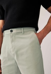 Pantalones ajustados verde claro en una tela suave y lisa, con un bolsillo frontal, presillas para cinturón y un detalle de botón oscuro.