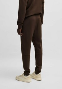 Bruine sweatpants van zacht materiaal met een taps toelopende pasvorm, voorzien van zijzakken en boorden bij de enkels, gecombineerd met crèmekleurige sneakers.