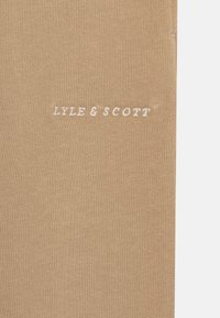 Beige szövet, bordázott textúrával, fehér színű "LYLE & SCOTT" hímzéssel. Egyszerű kialakítás, nincs további minta vagy díszítés.