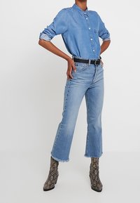 Denim shirt in light blue with button closure, rolled sleeves, paired with frayed hem, high-waist light blue jeans, and snake-print ankle boots.