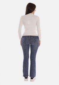 Top bianco a maniche lunghe e jeans svasati blu visti da dietro, con tasche posteriori visibili e dettagli di cucitura; il modello indossa sneakers bianche.