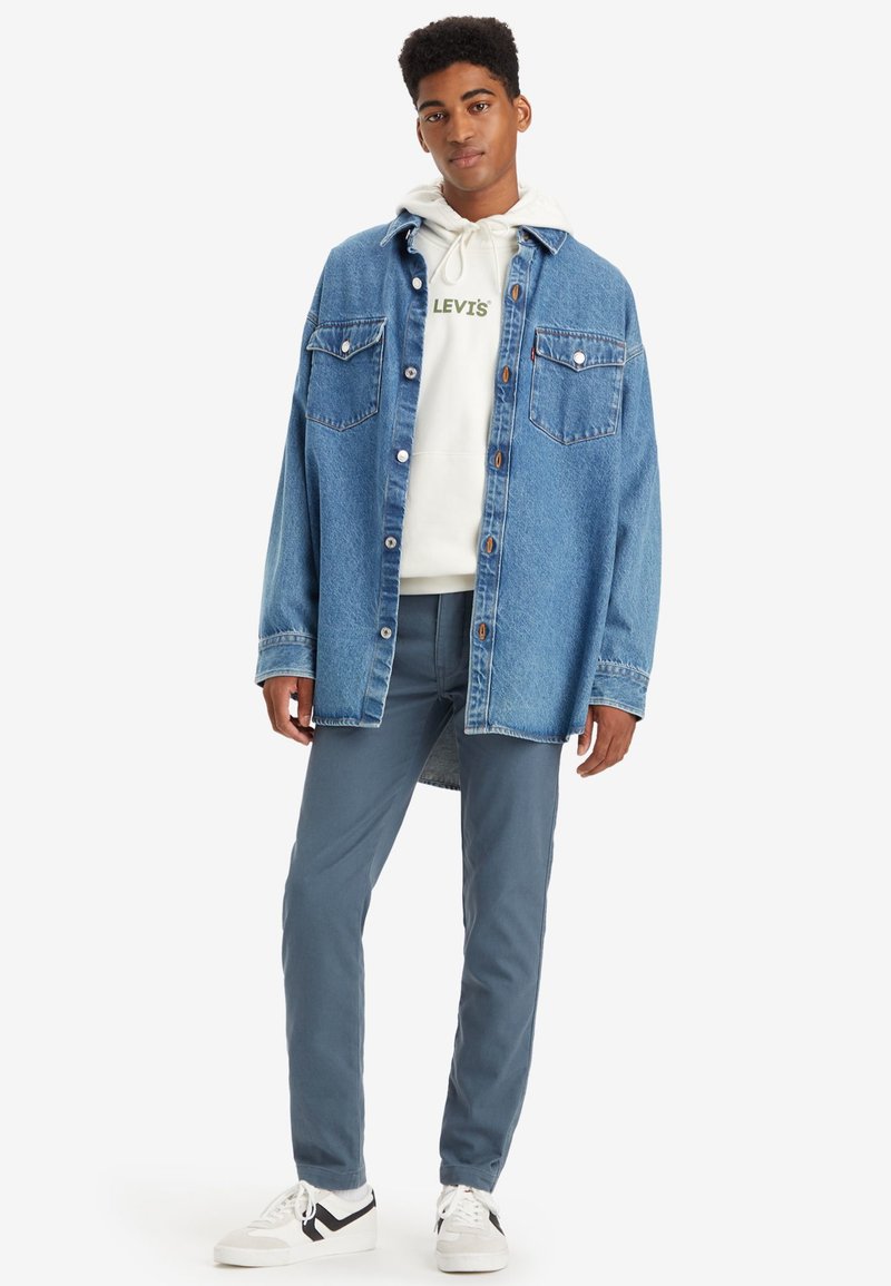 Chemise en jean bleu clair avec deux poches poitrine, fermeture à boutons, superposée sur un sweat à capuche blanc avec un logo vert, associée à un pantalon gris et des baskets.