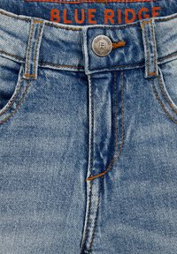 Džíny z denimu s světle modrým odstínem, oranžovým stehem, stříbrným knoflíkem s logem a štítkem s nápisem "BLUE RIDGE" v oranžové barvě.