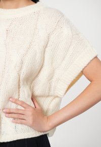 Strickpullover in Off-White mit Kabelmustern, kurzen Ärmeln und weit geschnitten. Weiche Textur mit gerippten Rändern.