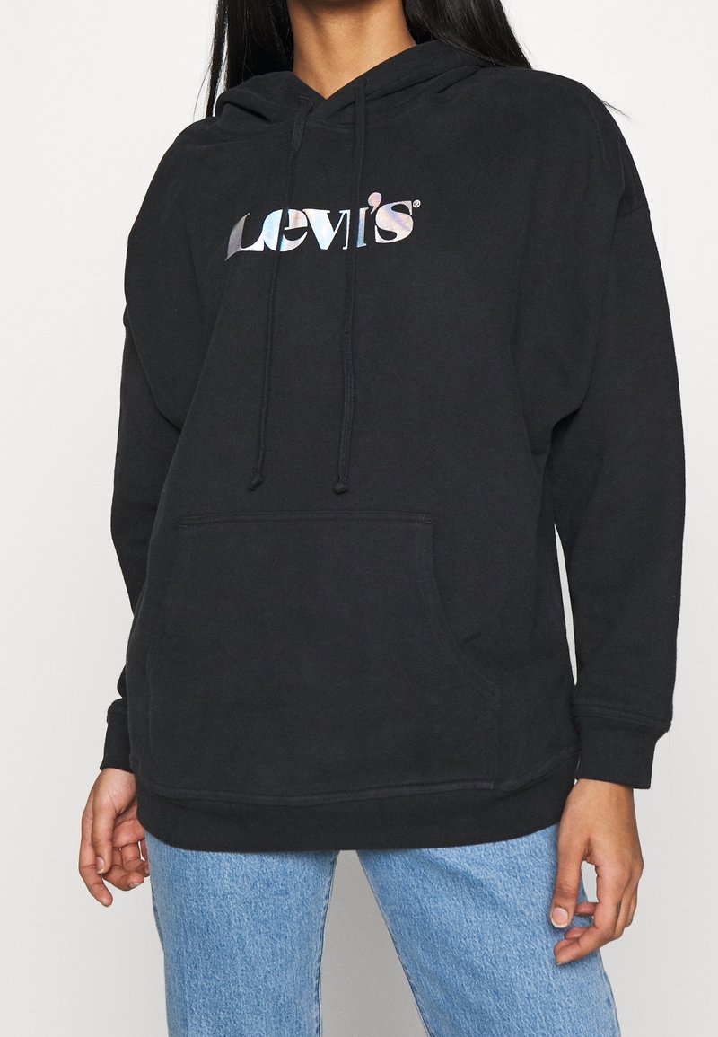 Personne portant un sweat à capuche noir Levi's avec logo holographique et poche avant, associé à un jean bleu clair, sur fond uni.