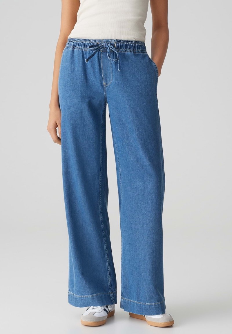 Personne portant un pantalon en jean bleu clair à jambe large avec taille à cordon, un débardeur blanc et des baskets blanches, mains dans les poches.